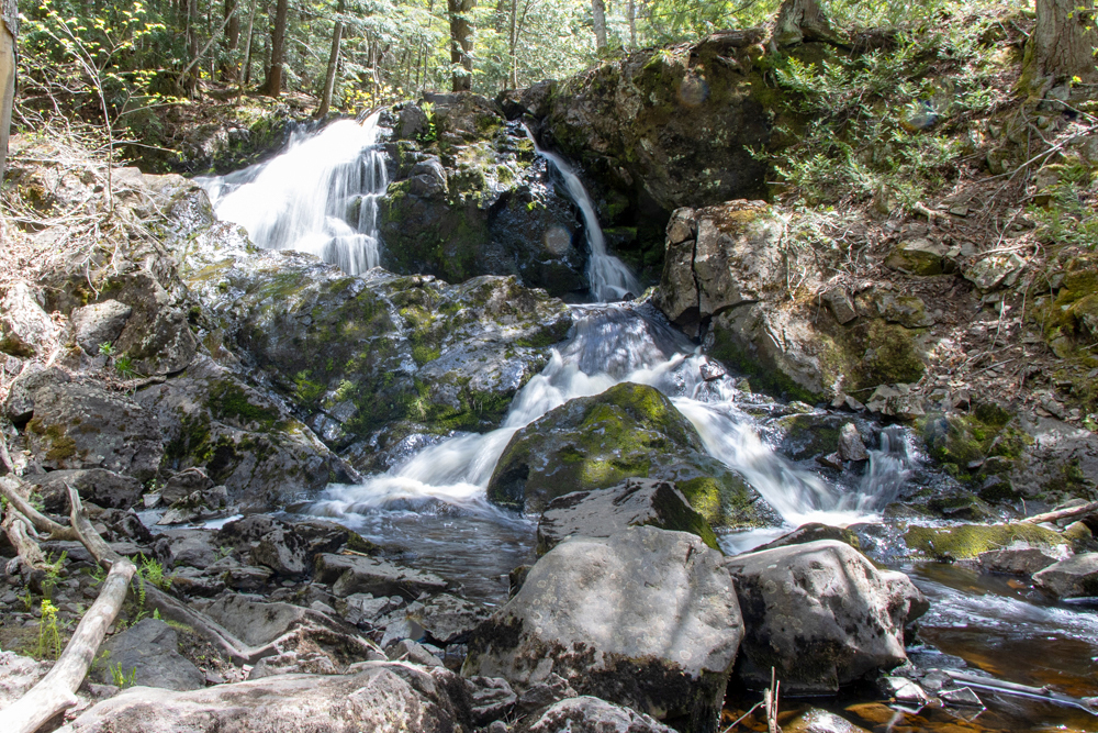 Long-exposure photograph of Wyandotte Falls, silky white water cascading over moss-covered rocks in a sunlit forest, photographed by Forbes Robertson in Michigan's Upper Peninsula.